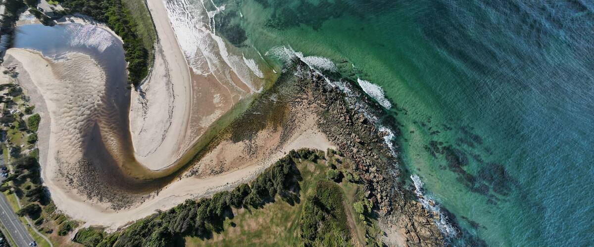 An aerial view of a coastal area with a sandy beach, rocky shoreline, and clear turquoise waters.