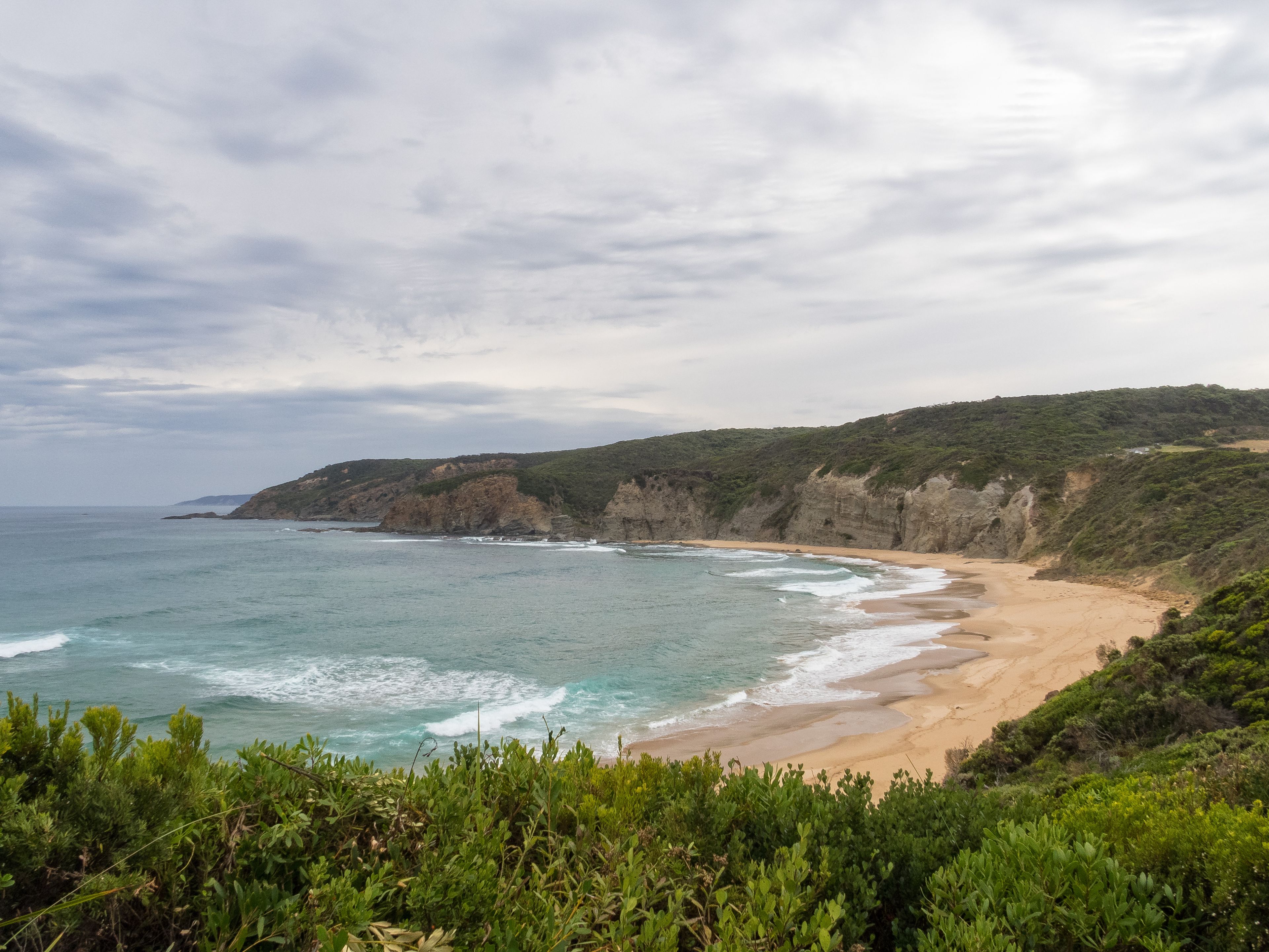 A coastal landscape with a sandy beach, rocky cliffs, and dense greenery.