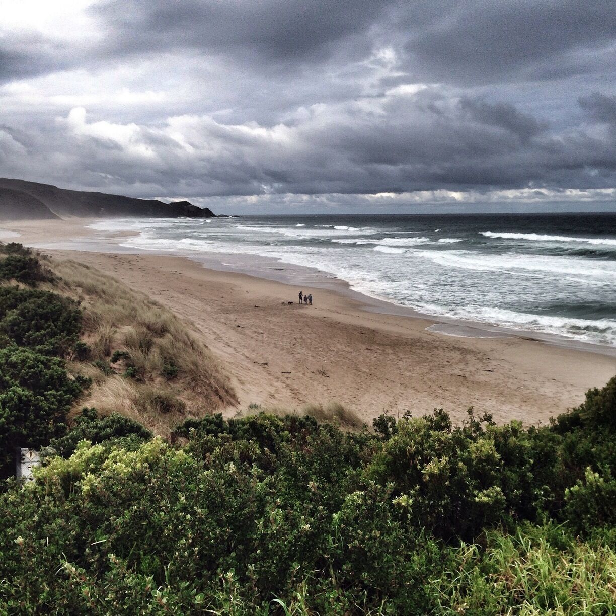 A beach with waves crashing onto the shore, a group of people standing on the sand, and a cloudy sky.
