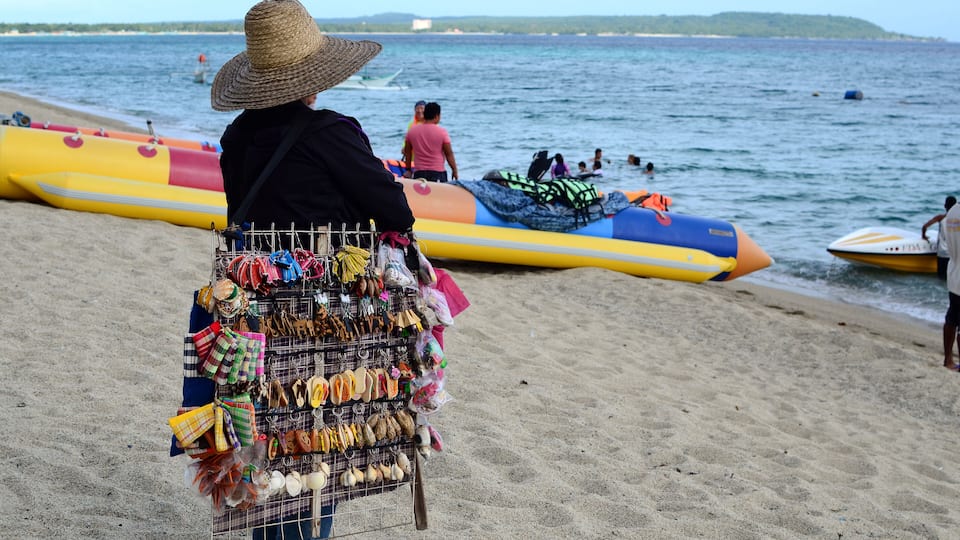 A beach with a vendor selling various items, including jewelry and bags, and inflatable boats in the water.
