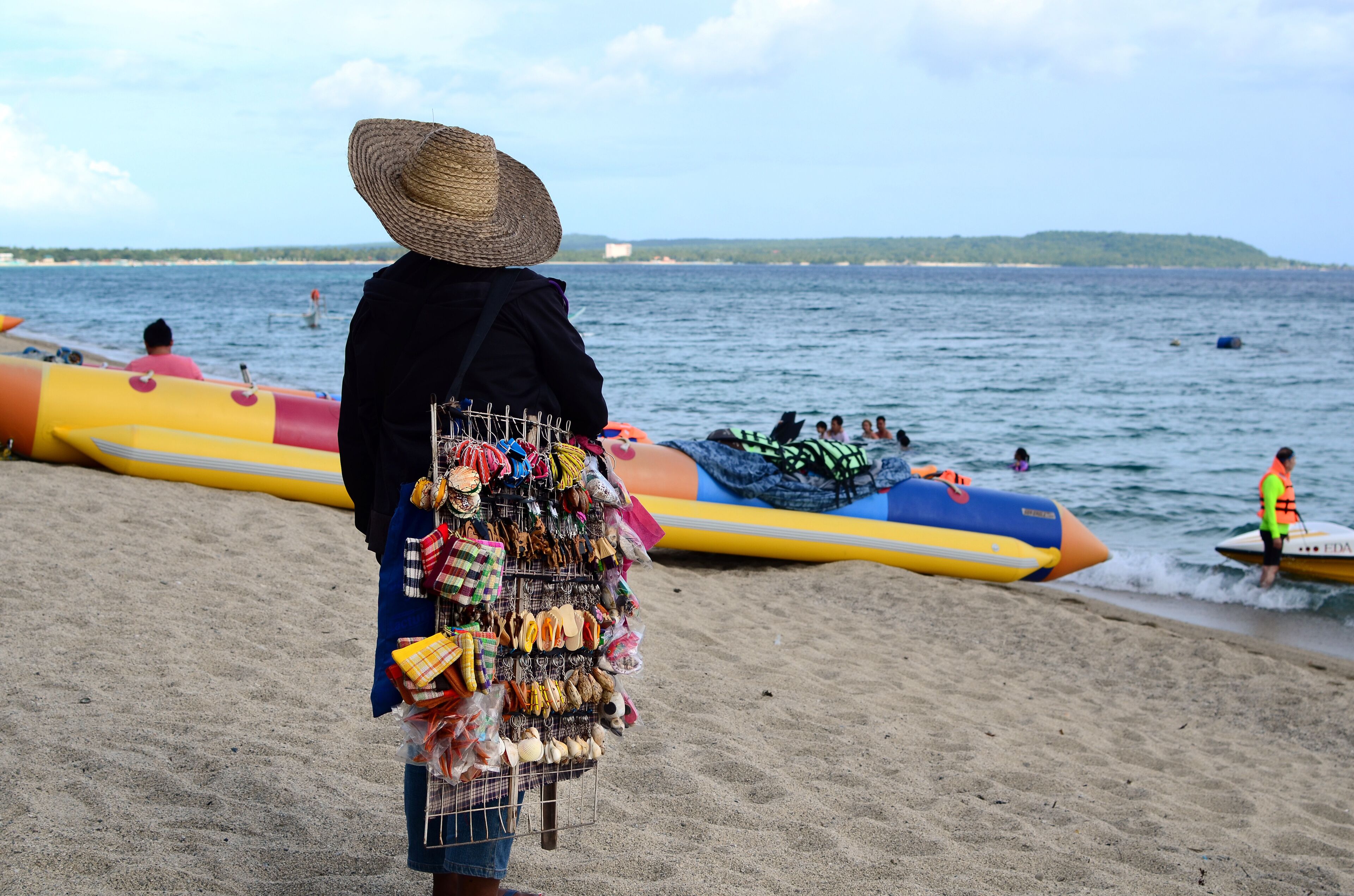 A beach with a person selling items, colorful inflatable boats, and a distant shoreline.