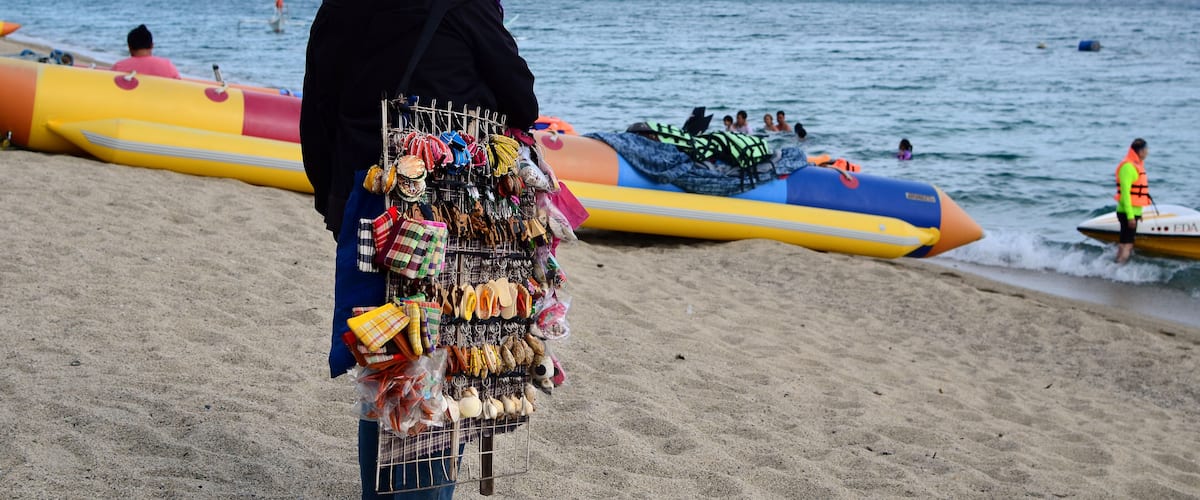 A beach with a person selling items, colorful inflatable boats, and a distant shoreline.