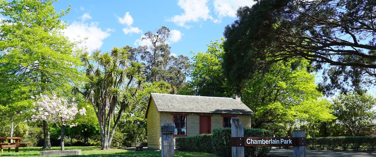 A park entrance with a sign reading "Chamberlain Park," surrounded by lush greenery and flowering plants.