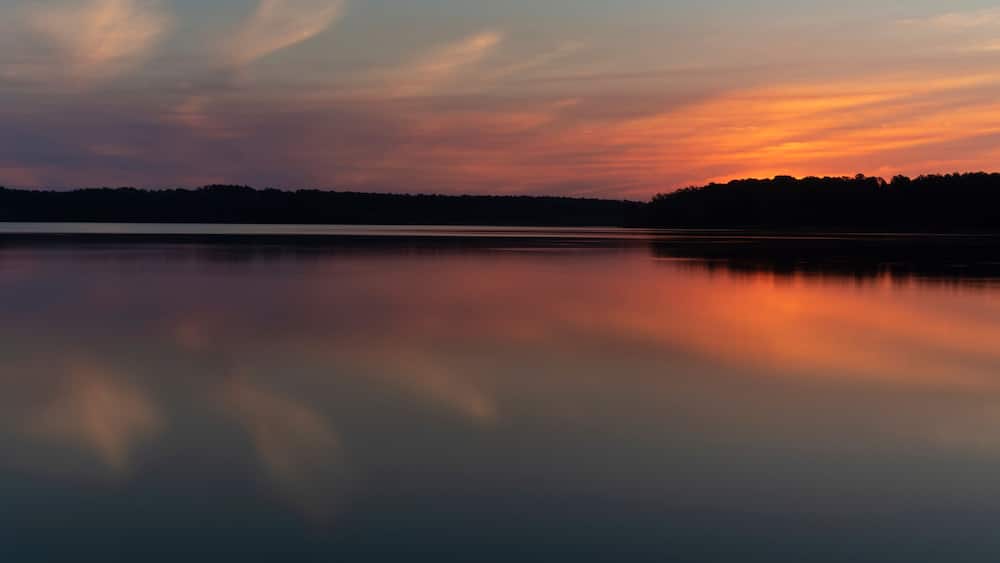 A calm lake at sunset with a silhouette of trees on the horizon.