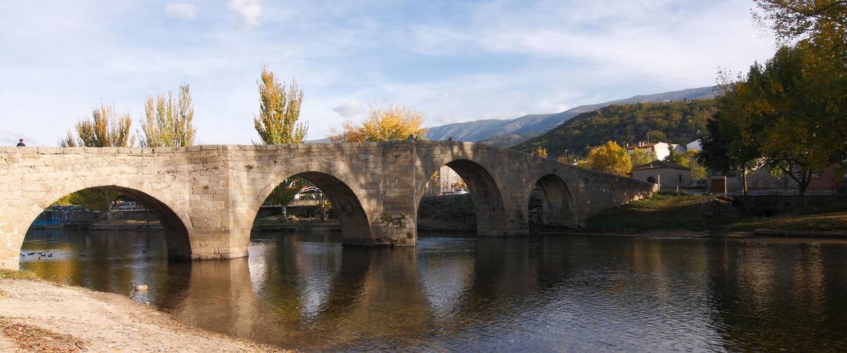 A stone bridge over a river with three arches, surrounded by trees and a hilly landscape.