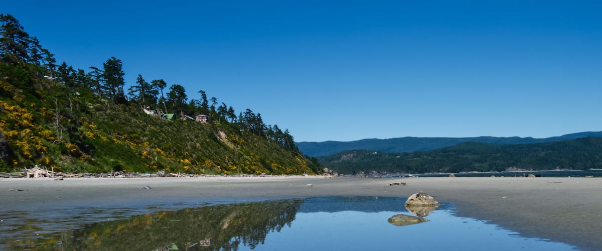 A beach with clear water reflecting the sky and surrounding landscape, a large rock in the center, and a forested hillside in the background.