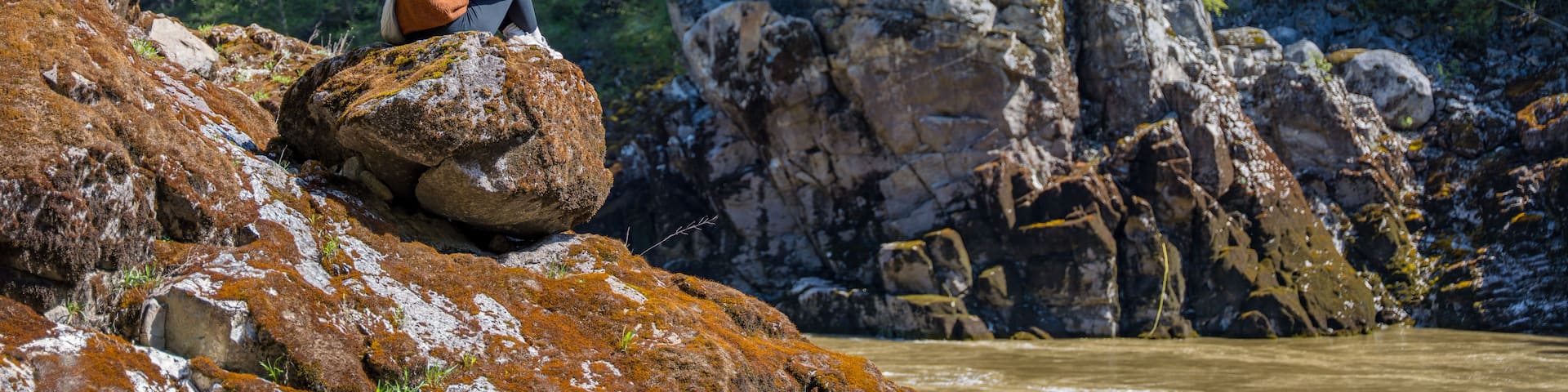 A person sitting on a rock by a river, a bridge, and a stone tower in a forested area.