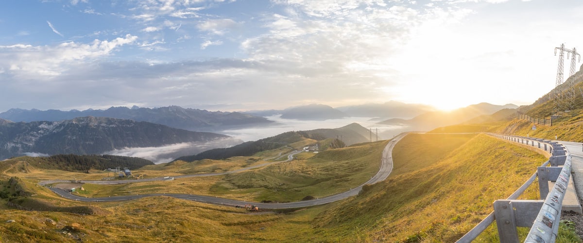 A winding mountain road with a guardrail, surrounded by lush green hills and distant mountains under a partly cloudy sky.