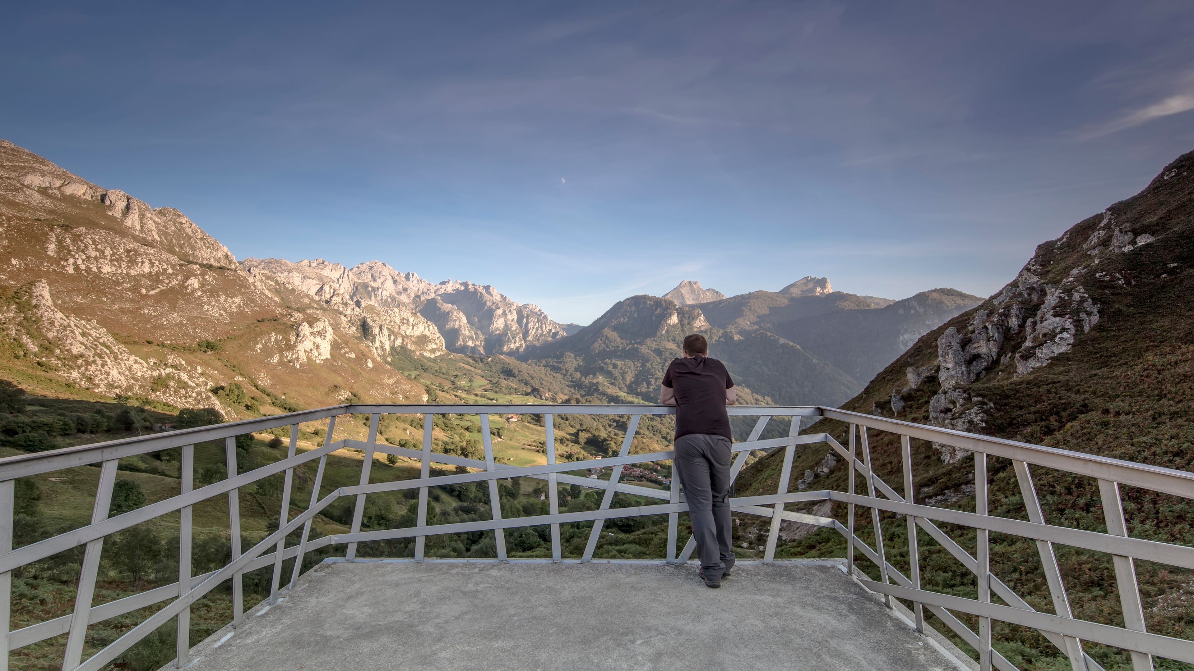 Une personne se tient sur une plateforme en béton, protégée par une rambarde métallique, et surplombe un paysage montagneux caractérisé par des falaises rocheuses et un ciel dégagé.