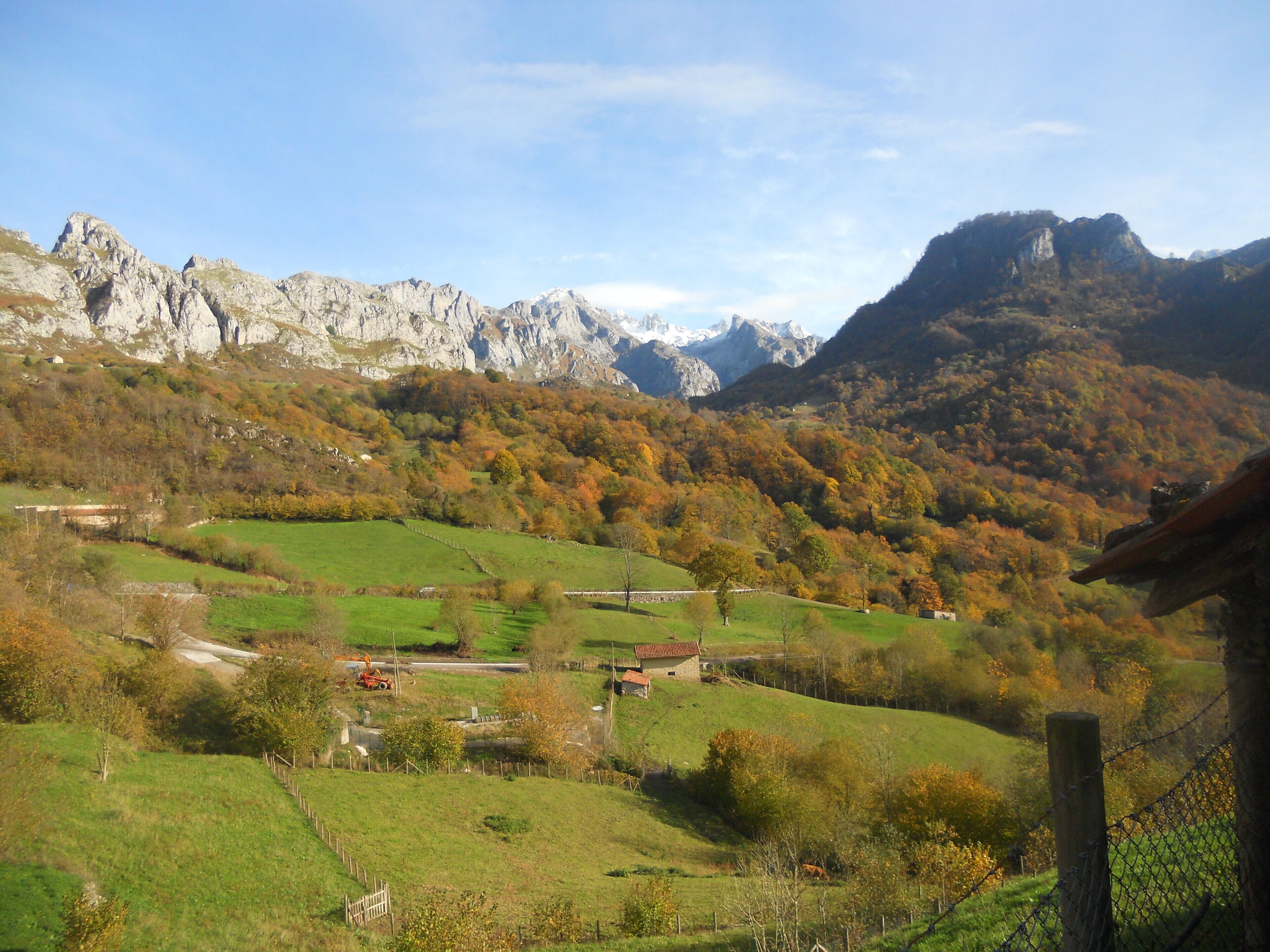 A mountainous landscape with autumn-colored trees, a green valley, and rocky cliffs.