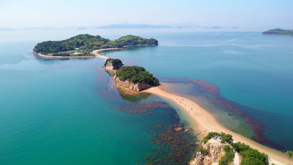 An aerial view of a coastal area with a sandy beach, clear turquoise waters, and green islands.