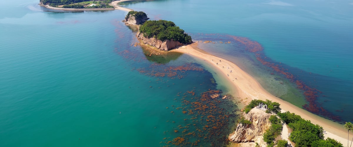 An aerial view of a coastal area with a sandy beach, clear turquoise waters, and green islands.