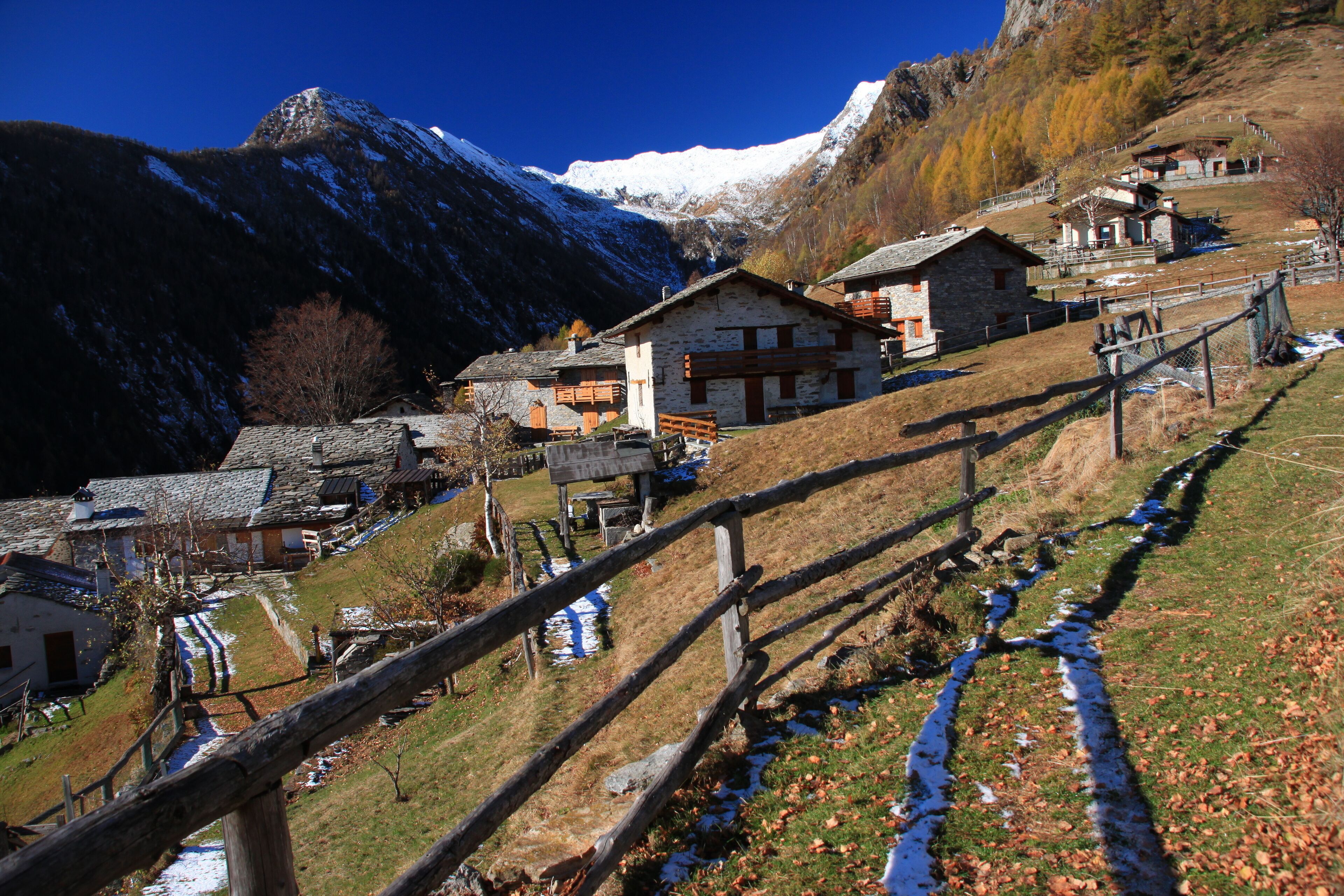A mountainous village with stone houses, a wooden fence, and a snow-capped peak in the background.