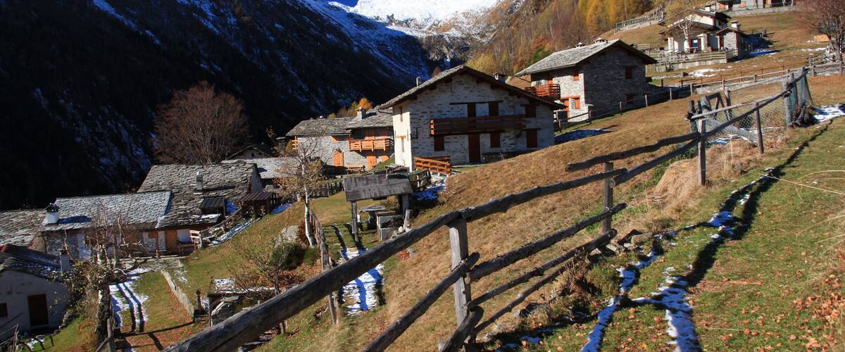 A mountainous village with stone houses, a wooden fence, and a snow-capped peak in the background.