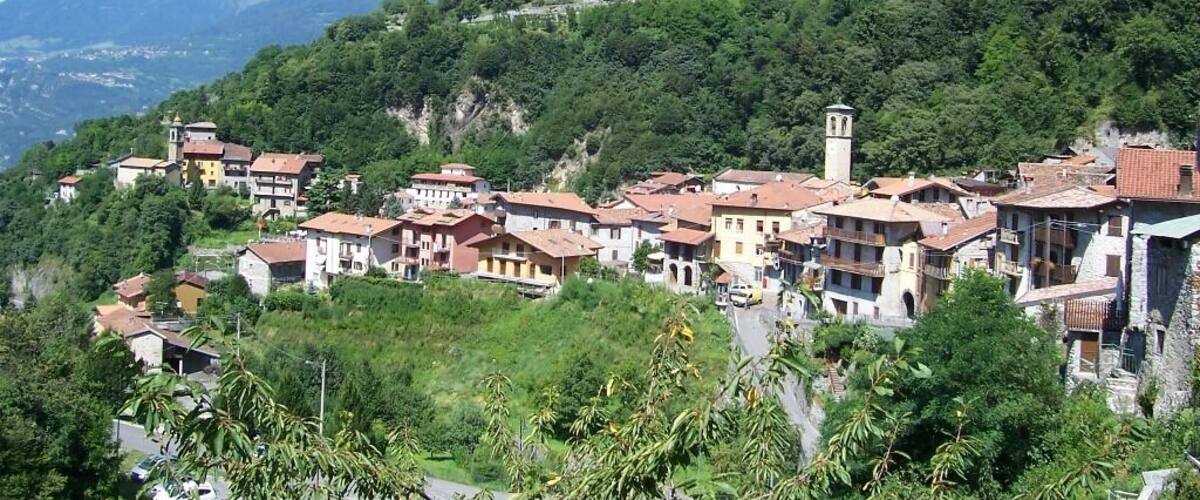 A hillside village with terracotta-roofed buildings, lush greenery, and a mountain in the background.