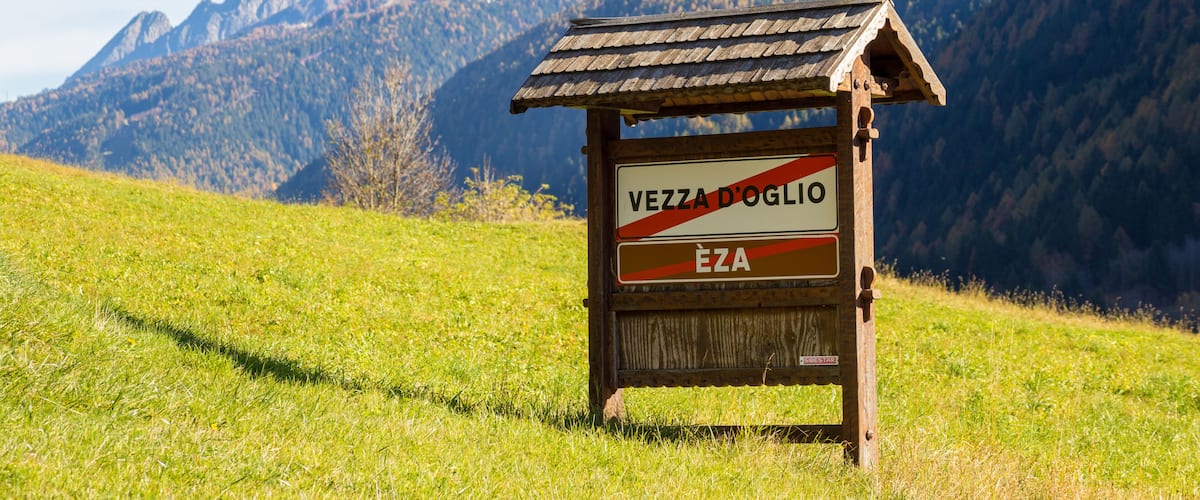 A wooden sign with 'VEZZA D'OGLIO' and 'ÈZA' written on it, set against a backdrop of mountains and a grassy field.