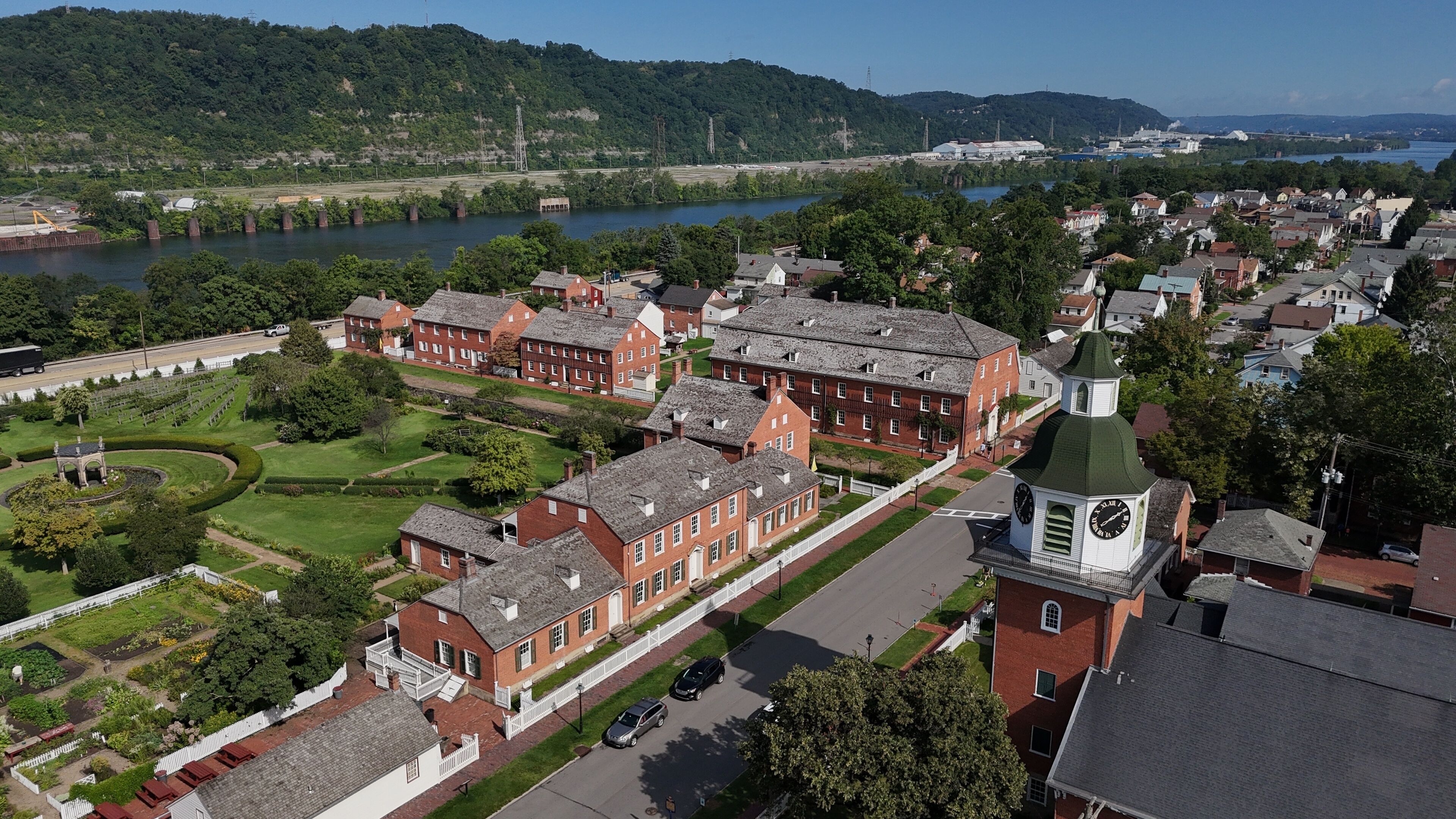 A historic red brick building complex with a clock tower, surrounded by lush greenery and a river.