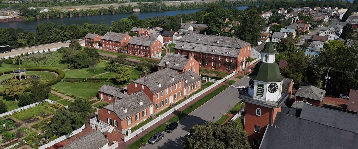 A historic red brick building complex with a clock tower, surrounded by lush greenery and a river.