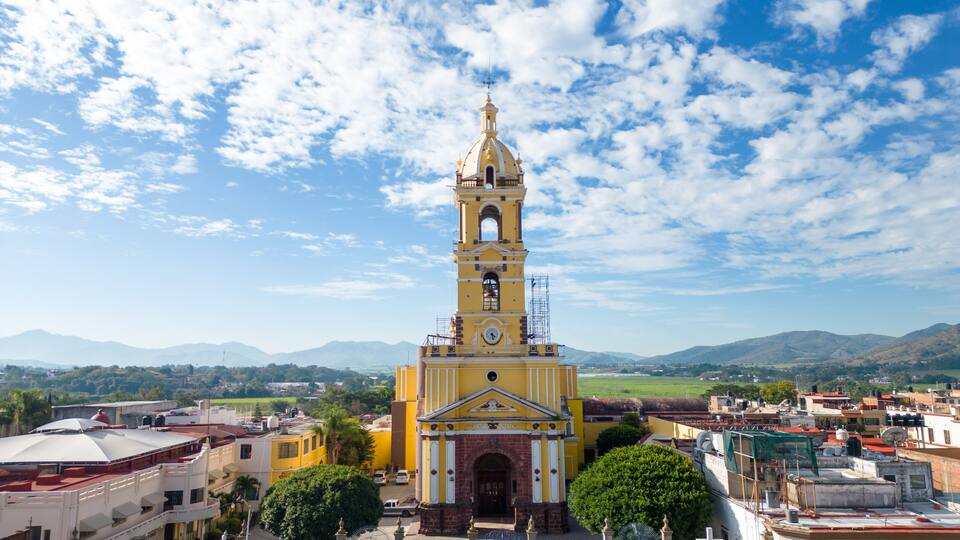 A yellow church with a bell tower, surrounded by buildings and mountains under a partly cloudy sky.