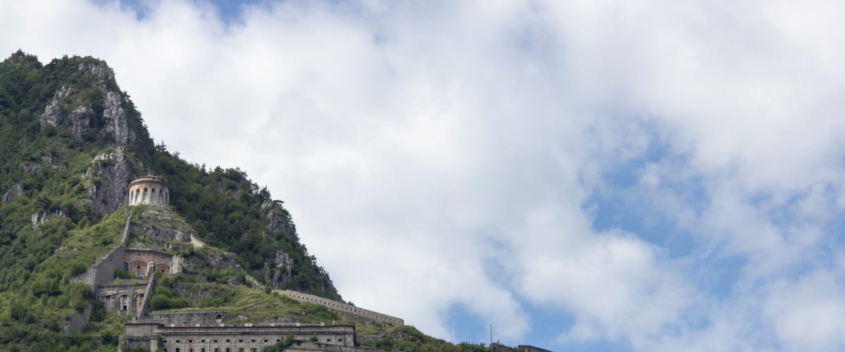 Une forteresse historique située sur une montagne, entourée d’une végétation luxuriante, sous un ciel partiellement nuageux.