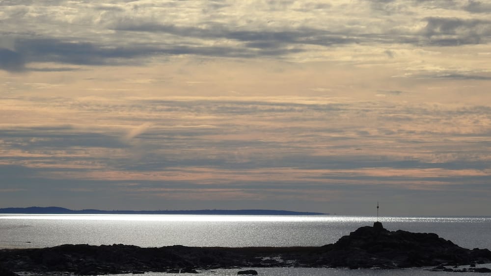 Une côte rocheuse parsemée de rochers qui émergent de l’eau, avec un phare visible au loin.