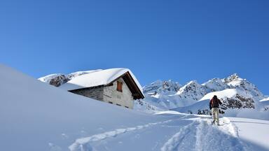 Paisaje nevado de montaña con un chalet de madera y un excursionista.