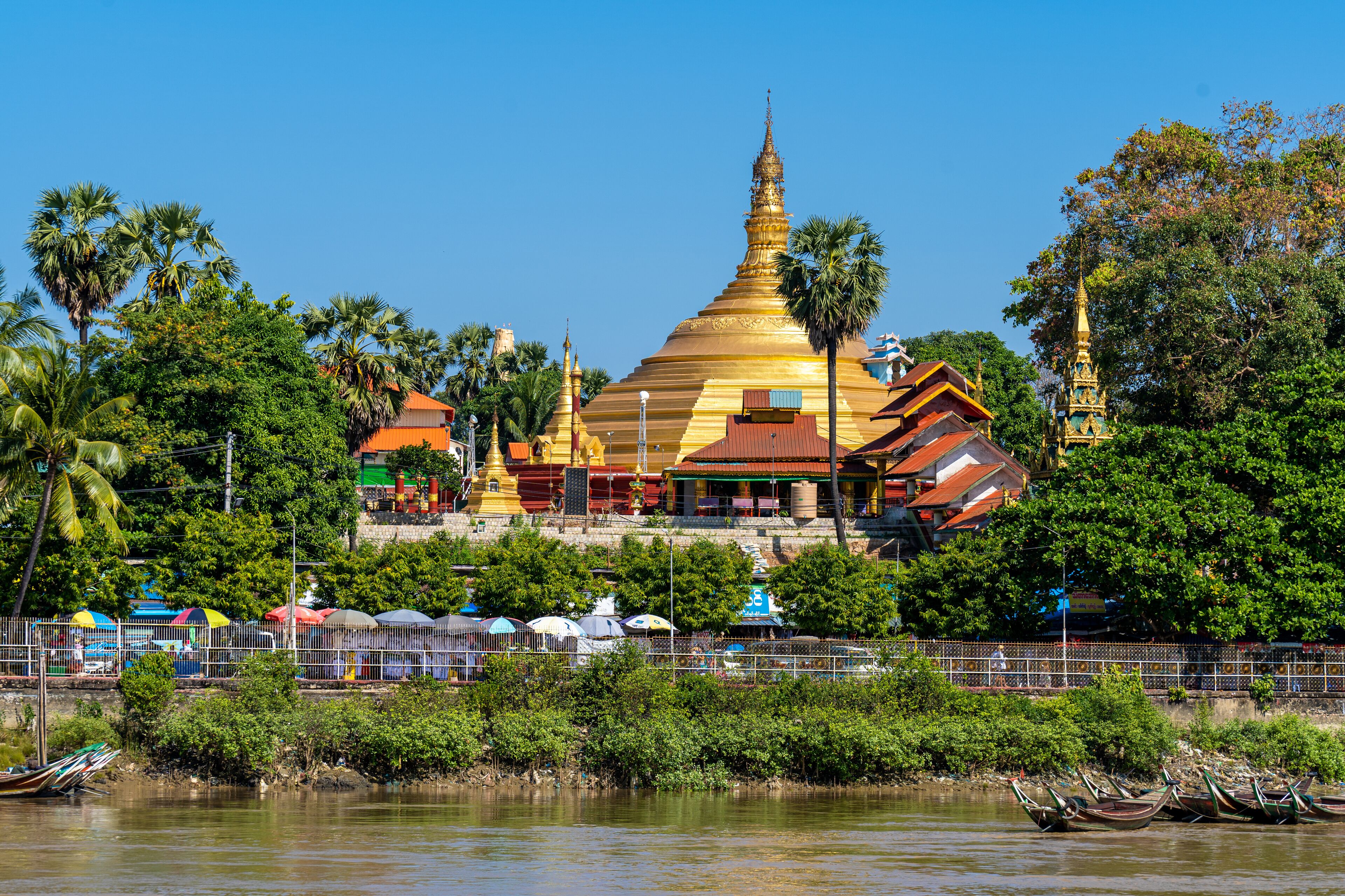 A golden pagoda surrounded by lush greenery and a river with boats.