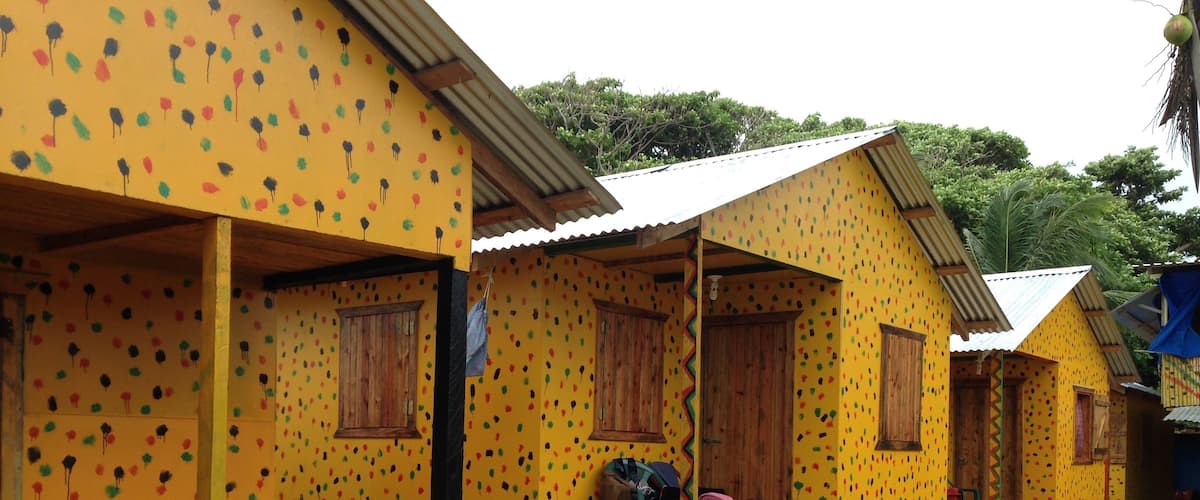 A row of colorful, yellow houses with wooden doors and windows, surrounded by greenery and a sandy ground.
