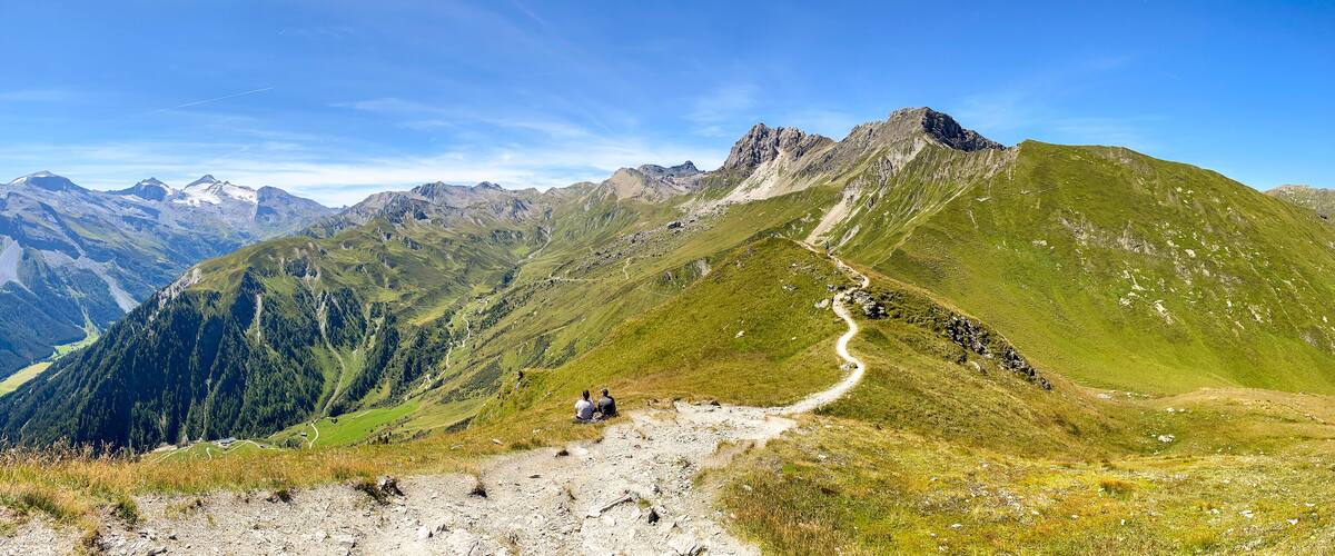 A mountain landscape with a winding path and two people sitting on a grassy area.