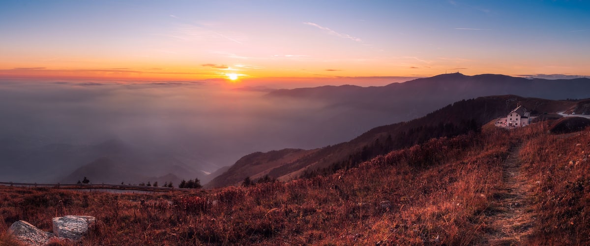 Un lever de soleil sur un paysage montagneux, avec un sentier menant à un bâtiment.
