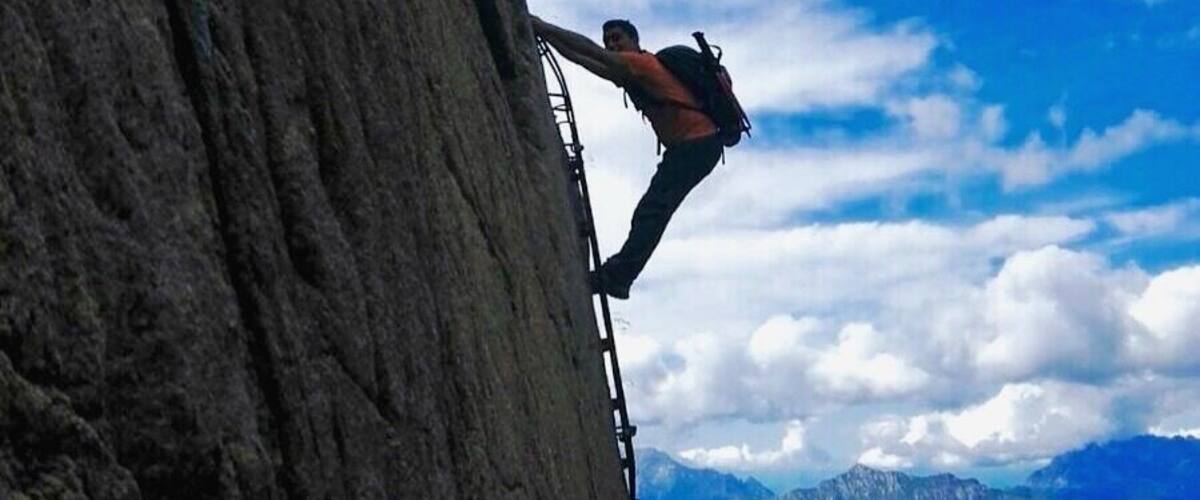 A person climbing a steep rock face with a mountainous landscape in the background.