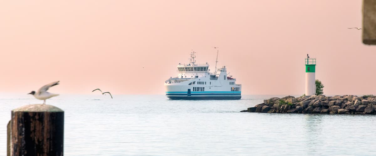 A ferry boat on calm waters, a lighthouse, and seagulls flying near the shore.