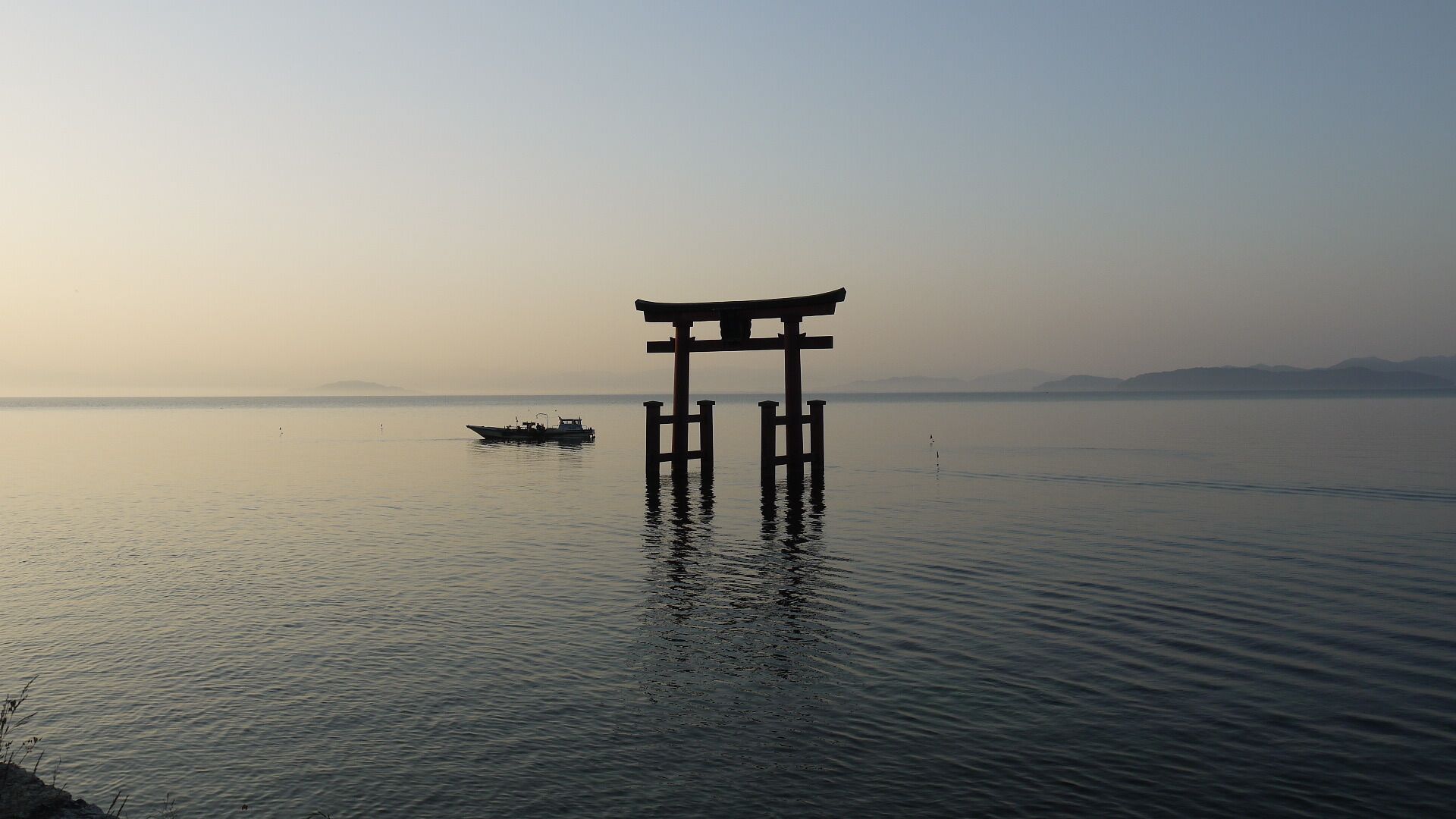 A calm body of water with a traditional Japanese torii gate in the foreground and a small boat in the distance.
