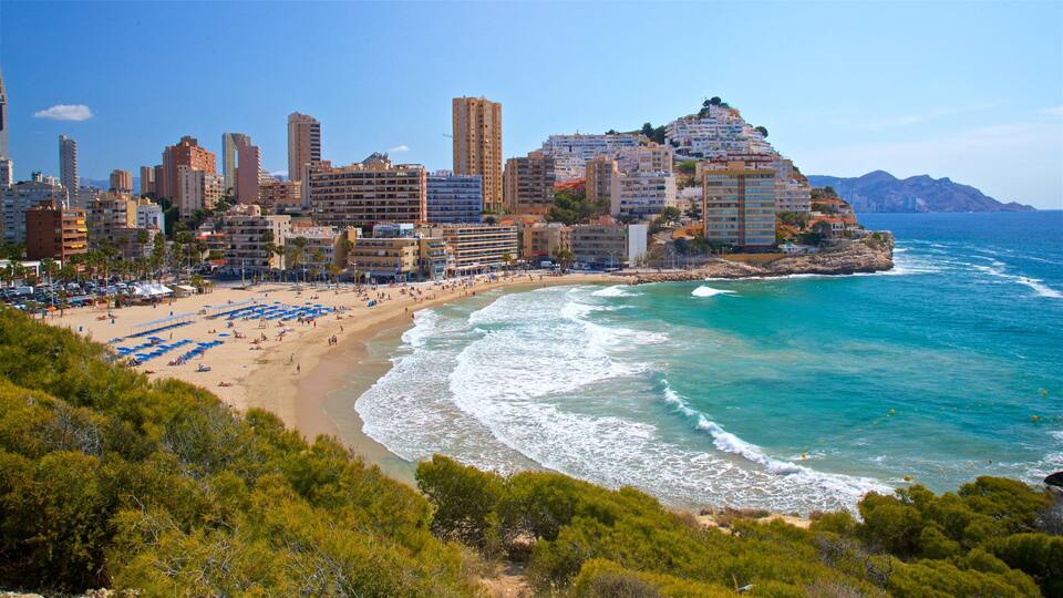 A coastal city with a beach, ocean waves, and a mountain with buildings on top.