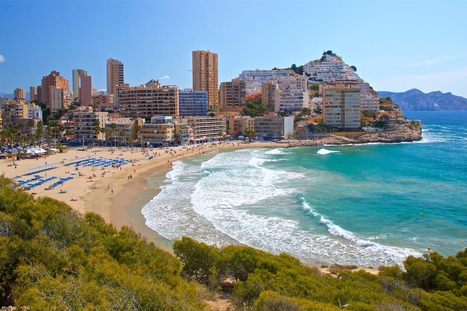 A coastal city with a beach, ocean waves, and a mountain with buildings on top.