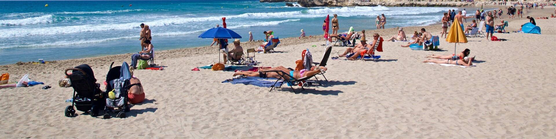 Una playa concurrida, gente tomando el sol, sombrillas y una colina rocosa al fondo.