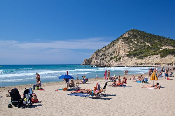Une plage bondée, où l’on voit des gens bronzer, des parasols et, au loin, une colline rocheuse.