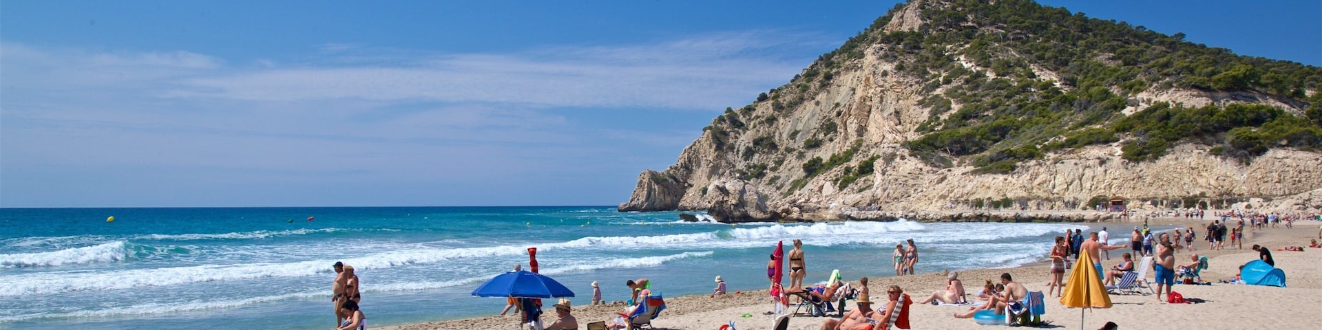 Una playa concurrida, gente tomando el sol, sombrillas y una colina rocosa al fondo.