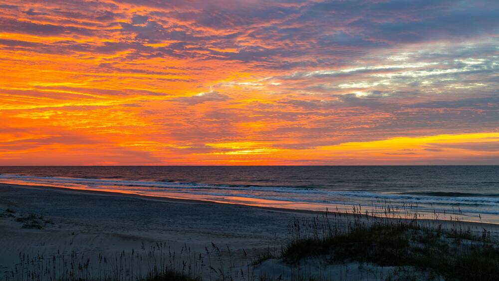A beach at sunset with vibrant orange and pink clouds, ocean waves, and sand dunes.