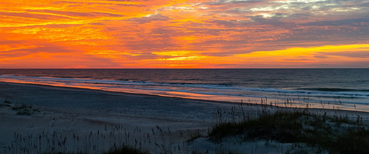 Ein Strand bei Sonnenuntergang mit leuchtend orangefarbenen und rosafarbenen Wolken, Meereswellen und Sanddünen.