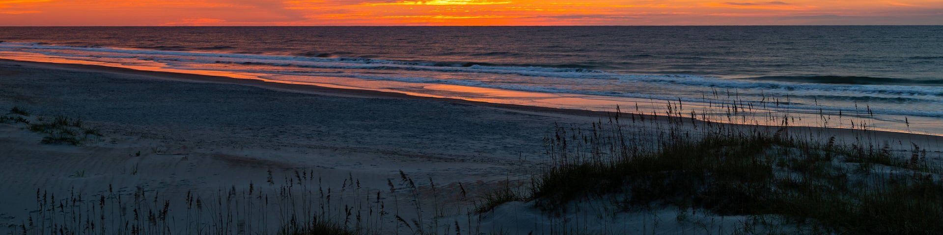 A beach at sunset with vibrant orange and pink clouds, ocean waves, and sand dunes.