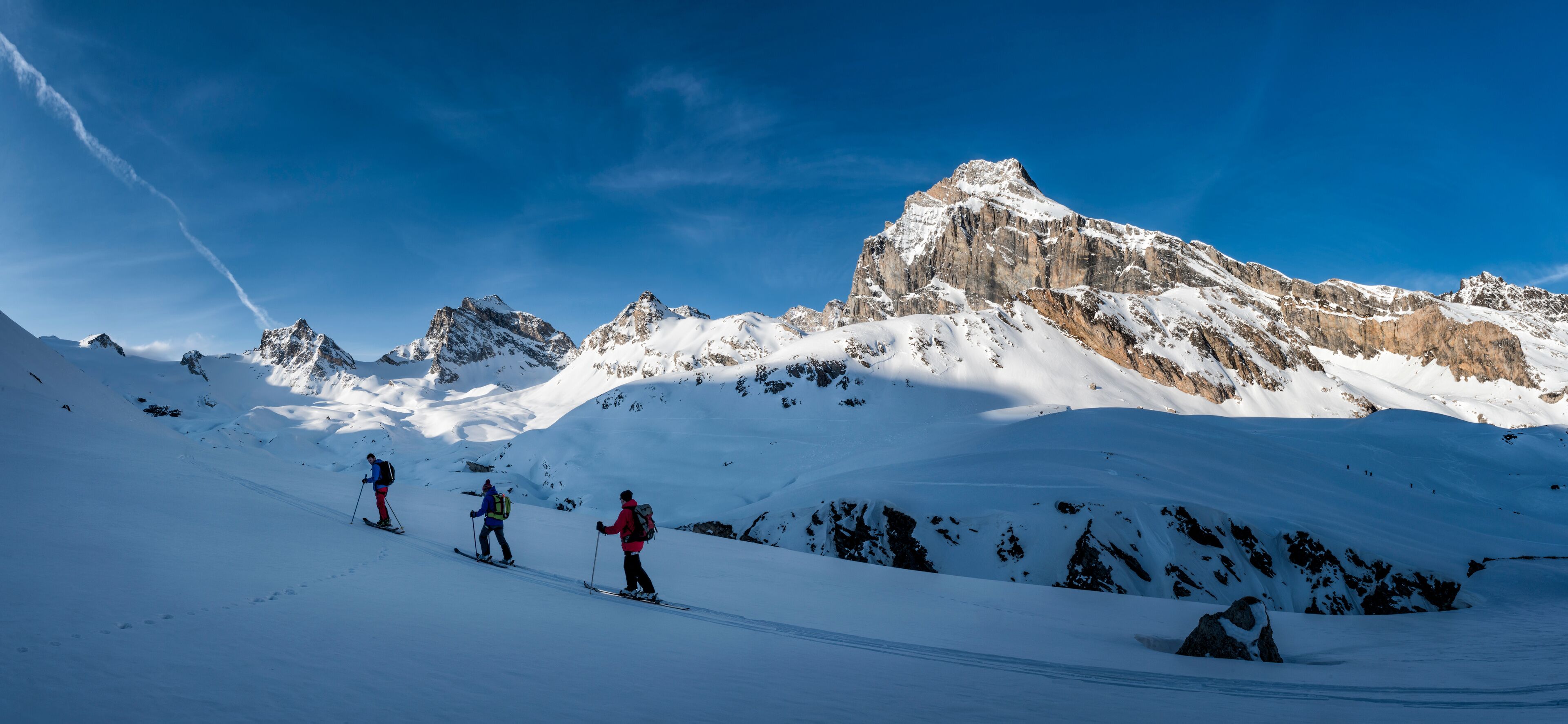 A group of skiers on a snow-covered mountain slope with rocky peaks in the background.