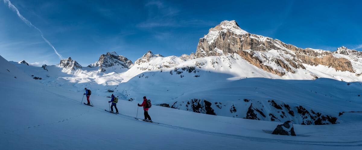A group of skiers on a snow-covered mountain slope with rocky peaks in the background.
