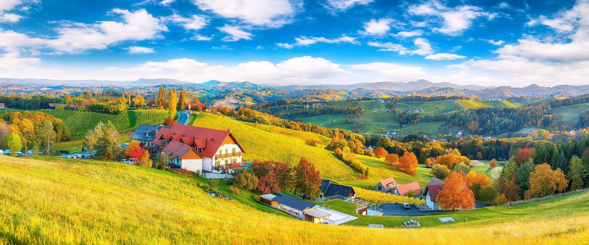 A rural landscape with rolling hills, a red-roofed house, and autumn-colored trees.