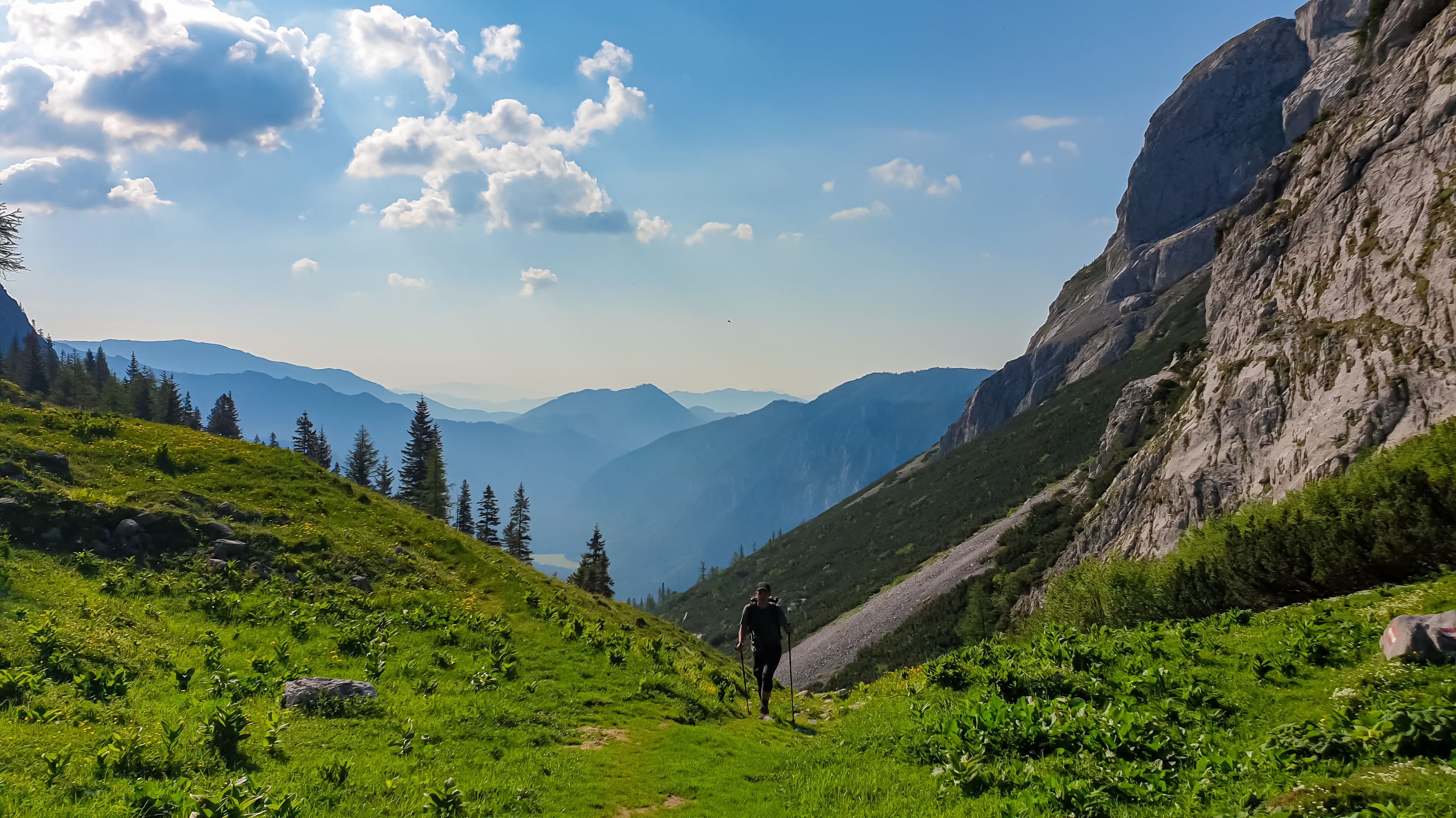 Een wandelaar op een begroeiing bedekt bergpad, met een rotsachtige klif en bergen in de verte.