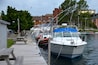 En bro med flere både fortøjet langs siden, en træbrygge og bygninger i baggrunden.