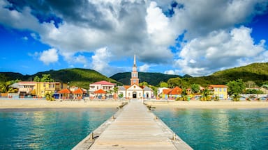 A coastal town with a wooden pier leading to a church with a tall steeple, surrounded by colorful buildings and a hilly backdrop.