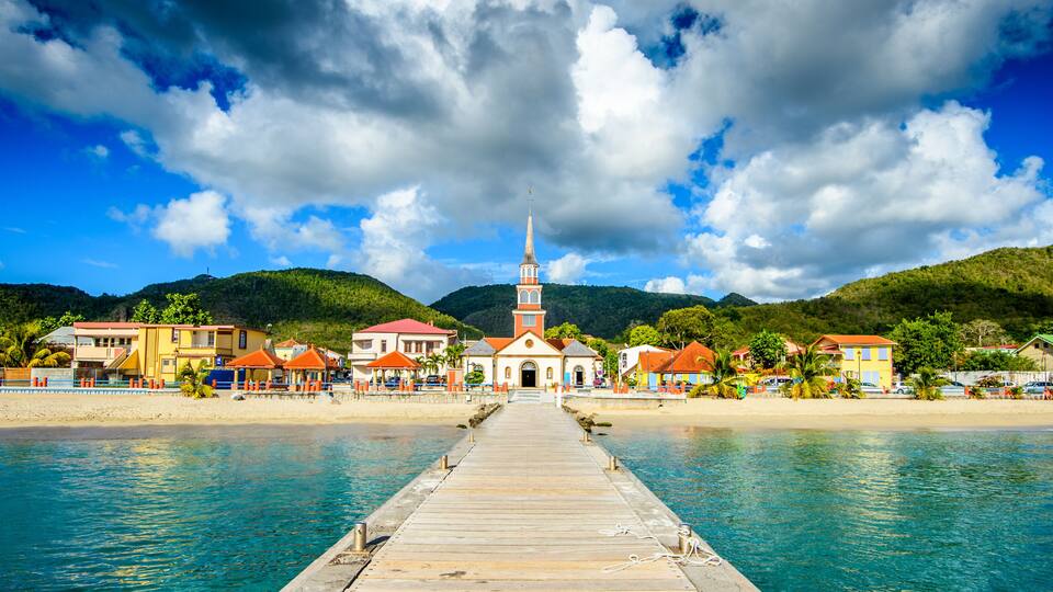 A coastal town with a wooden pier leading to a church with a tall steeple, surrounded by colorful buildings and a hilly backdrop.
