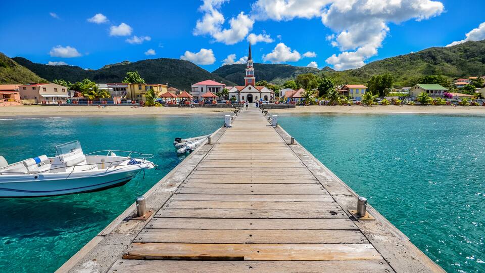 A wooden pier leading to a small town with a church, boats, and clear turquoise water.