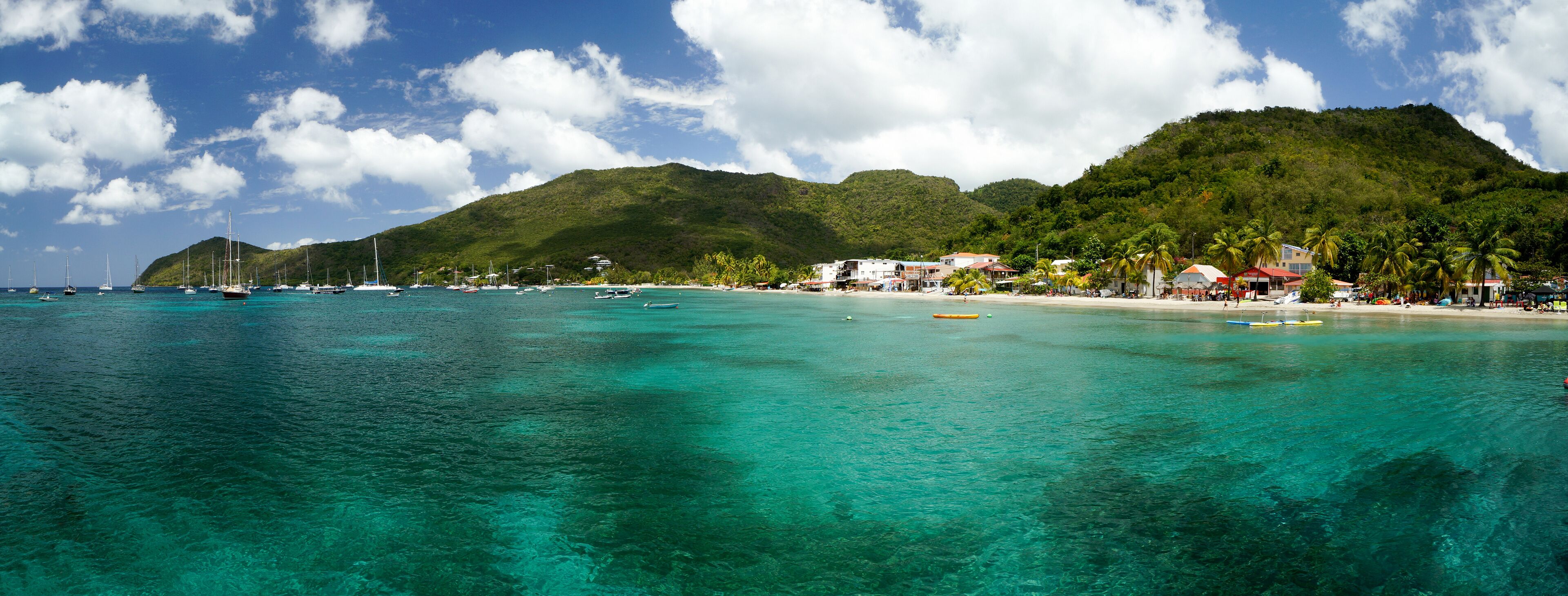 A tropical beach with clear turquoise waters, a small town with buildings and palm trees, and a mountain in the background.